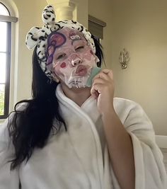 a woman in a white robe is holding up a mask with her face painted pink and black