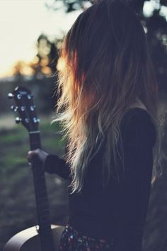 a woman with long hair holding a guitar in her hand and looking at the ground