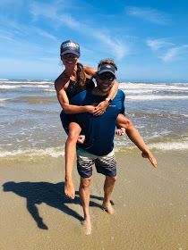 a man carrying a woman on his back in the sand at the beach with water behind him