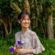 a woman holding a purple flower in her hand and smiling at the camera while standing next to some trees