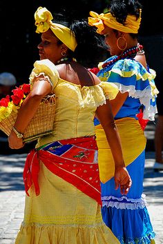 two women in colorful dresses are walking down the street with baskets on their heads and one is carrying flowers