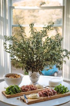 a table topped with plates and bowls next to a vase filled with greenery on top of a wooden cutting board