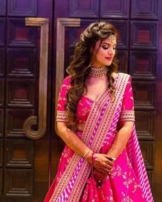 a woman in a pink lehenga standing by a wooden door with her hands on her hips