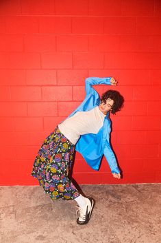 a woman is posing in front of a red wall wearing a blue jacket and floral skirt