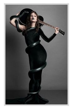 Taylor Swift wearing a long one-sleeved black body con dress holding a black guitar over her head with a long black snake wrapped around her body