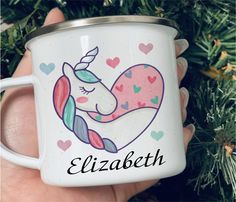 a hand holding a white coffee mug with a unicorn on it's face and hearts