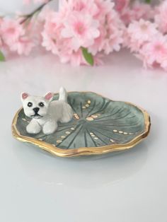 a small white dog sitting on top of a green leaf shaped dish with pink flowers in the background