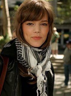 a woman in black jacket and white scarf standing on the street with her head turned to the side