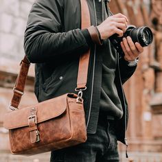 a man holding a camera in his right hand and wearing a brown leather messenger bag