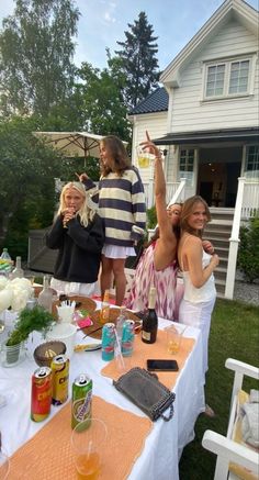 three women standing around a table with food and drinks on it, in front of a house