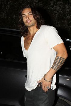 a man with long hair and tattoos standing in front of a black car at night