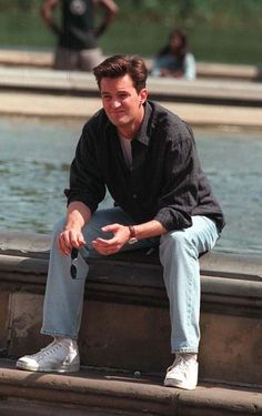 a man sitting on the edge of a fountain with his hands in his pockets and smiling