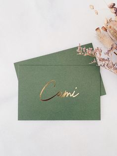 two green envelopes with gold foil lettering on them and some dried flowers in the background