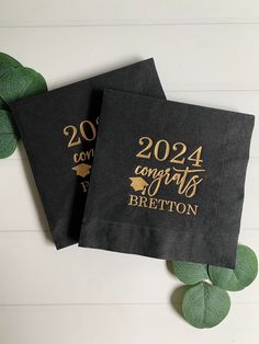 two black graduation napkins with gold foil lettering on them and green leaves around the edges