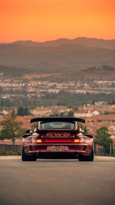 the back end of a red sports car parked on top of a parking lot with mountains in the background