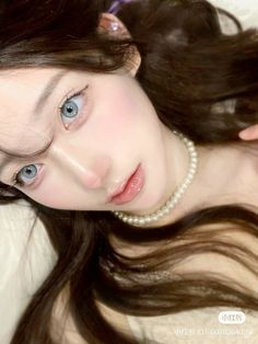 a woman with long brown hair and blue eyes laying on a white bed wearing a pearl necklace