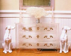 two white dogs sitting on the floor in front of a dresser with flowers and a mirror