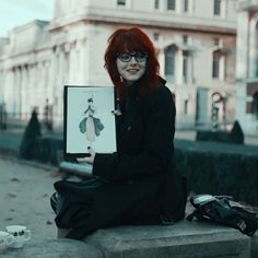 a woman with red hair and glasses sitting on the ground holding up a piece of paper