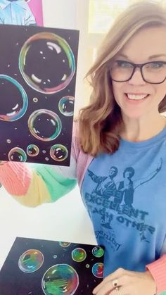 a woman in glasses holding up some art work with soap bubbles on the paper and water droplets