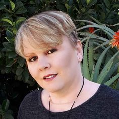 a woman with blonde hair wearing a black shirt and necklace standing in front of some plants