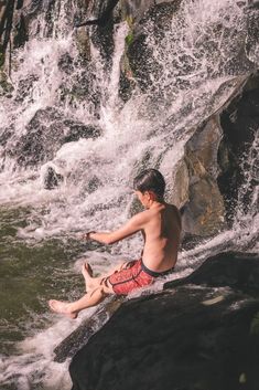 a young man sitting on top of a rock next to a body of water near a waterfall