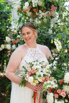 a woman in a white dress is holding a bouquet and smiling at the camera while standing under a floral arch