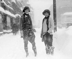 two women standing in the snow next to a street sign and building with lots of snow on it