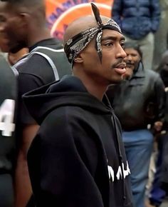 a man wearing a bandana standing in front of other men on the street with people behind him