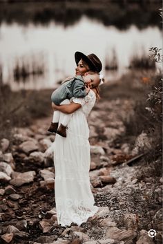 a woman holding a baby in her arms while standing on rocks near the water's edge