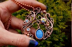 This design features a most unique gem setting technique showing off a stunning, blue opal is October's birth stones and three diamond cut topaz which are December's birth stone. The gems are set in pure, antiqued copper in an intricate design. The piece is completed with a mystic CZ as an accent. Chain is included Size is:  50 mm tall by 50 mm wide.  The item will be by sent tracked and signed post without a gift box to keep the shipping cost down. If you would like a gift box or other shipping upgrades select the desired option at check out. For free videos and new tutorial releases (paid and free), exclusive discount codes and promos sign up to my newsletter, follow on my Facebook, Instagram or YouTube channel: Craft community & support group: https://www.facebook.com/groups/wirewrapart Copper Gemstone Pendant Jewelry, Unique Wire Wrapped Round Jewelry, Handmade Opal Necklaces As Gift, Handmade Opal Necklaces For Gift, Handmade Opal Pendant Necklace, Fusion Style Wire Wrapped Pendant Jewelry, Handmade Elegant Opal Necklace, Copper Wire Necklace With Oval Pendant As Gift, Elegant Gemstone Copper Jewelry