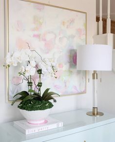 a white vase with flowers on top of a table next to a lamp and painting