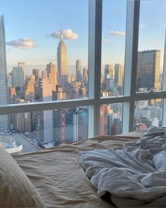 an unmade bed in front of a large window with cityscape and skyscrapers