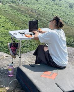 a woman sitting at a table with a laptop on top of a rock overlooking a valley