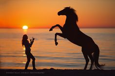 a woman standing next to a horse on top of a beach near the ocean at sunset