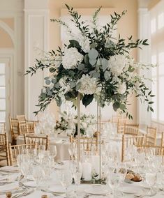 a tall vase filled with white flowers and greenery on top of a dining room table