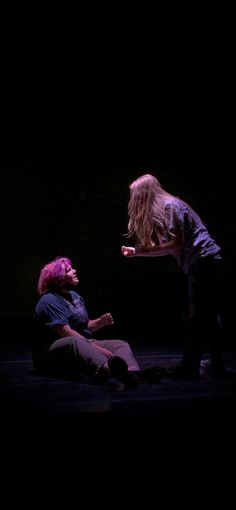 two people sitting on the ground in front of a black background, one person with pink hair