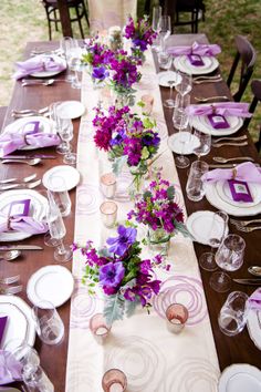 a long table is set with purple flowers and place settings for the guests to sit at