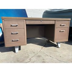 a brown desk with three drawers and two wheels on the bottom, in front of a blue car