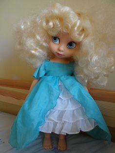 a doll with blonde hair wearing a blue dress and sandals on a bed in a room