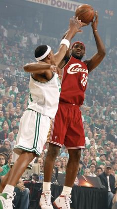 two men playing basketball in front of an audience at a sporting event, one is jumping up to grab the ball