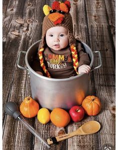 a baby wearing a turkey hat sitting in a pot with apples and oranges next to it