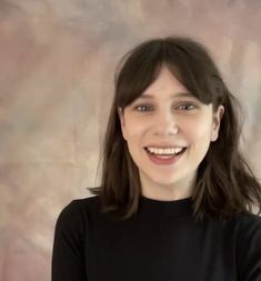 a woman with long hair smiling at the camera and wearing a black shirt, standing in front of a wall