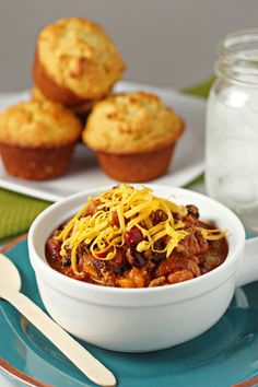 a white bowl filled with chili and cheese next to muffins