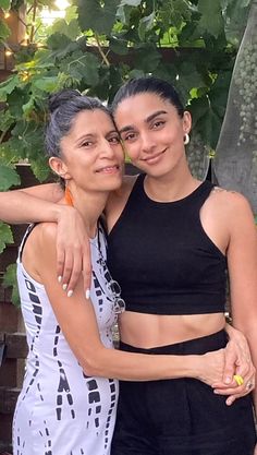 two women standing next to each other in front of a tree and some bushes with their arms around one another