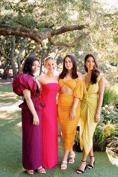 three women standing next to each other in front of trees and grass with one woman wearing an orange dress