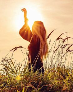 a woman is standing in tall grass with her arms up and the sun shining behind her