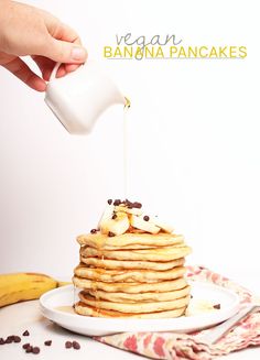 a person pouring syrup onto a stack of pancakes with chocolate chips and bananas on the side