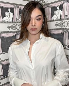 a woman standing in front of a wallpapered background wearing a white shirt and diamond earrings