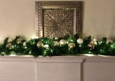 a fireplace mantel decorated with green and white christmas garland, ornaments and lights on the mantle