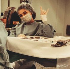 a woman sitting at a table with her hands in the air while wearing a surgical mask